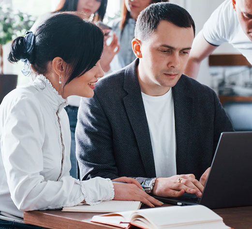 A focused man and woman looking at a laptop, possibly discussing book editing services with colleagues in the background.